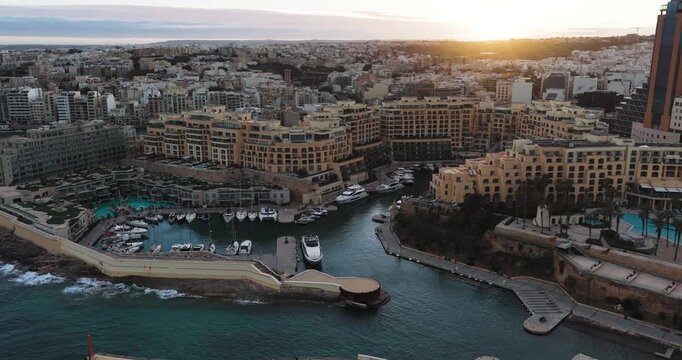 Aerial view of Portomaso Marina with luxury yachts docked in the harbor alongside elegant buildings, reflecting the golden light, St. Julian, Malta.