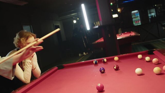 Refined caucasian woman carefully prepares for shot on red bright pool table while her friend observes nearby. A blonde female player places her hand in a bridge position to hold a cue at vintage club