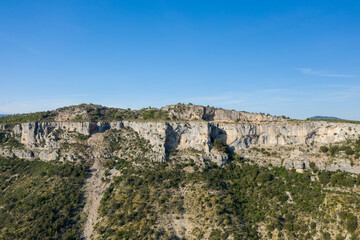 Fototapeta premium Steep limestone escarpment rises above a densely vegetated hillside at Cirque de Navacelles. Bright sunlight and clear blue sky highlight the rocky textures and natural contours.