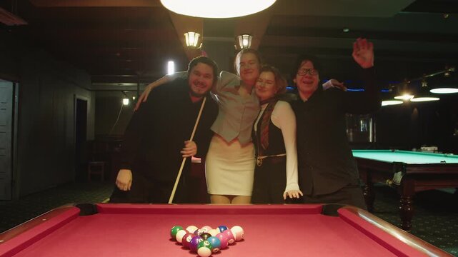 Friends celebrate with laughter. Caucasian men and women cheer and pose around red billiards table with balls. Four companions smile, hug and raise arms during lively night at bar in casual atmoshere