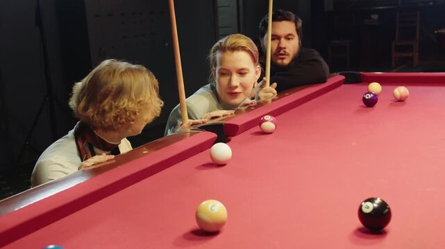 Casual gathering of friends engaging in friendly pool match. Group of caucasian youthful man and women with cue are discussing and joking leaning on the red table while playing billiards in night club