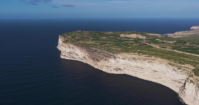 Aerial view of imposing, layered cliffs juxtapose the deep blue ocean, a rugged coastline, and lush greenery above, Xlendi, Munxar, Malta.