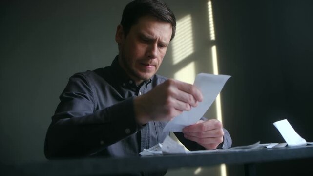Worried man sitting at table meticulously checking receipts, calculating expenses, feeling stress financial hardship, inflation, budget management in home. Man reviewing household bills, home expenses