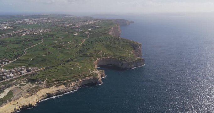 Aerial view of the rugged Xlendi cliffs meeting the turquoise sea, contrasting against the green terraced fields and coastal town, Xlendi, Munxar, Malta.