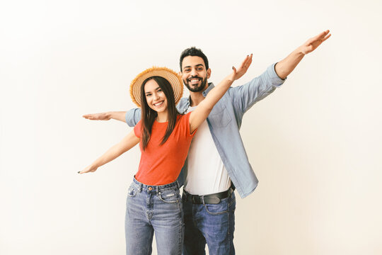 Young couple in love pretending airplane flight, spreading hands like wings, having fun while posing over light studio wall, looking and smiling at camera