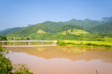 Obraz premium A modern bridge spans a wide muddy river, reflecting lush green hills and a clear blue sky in northern Vietnam. The tranquil landscape features vibrant fields, distant mountains, and bright summer