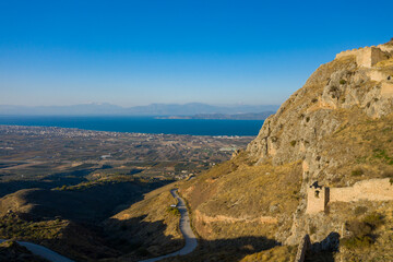 Fototapeta premium Golden rocky slopes of Acrocorinth with ancient stone walls descend toward a vast plain and the shimmering Corinthian Gulf under a clear blue sky in Greece.