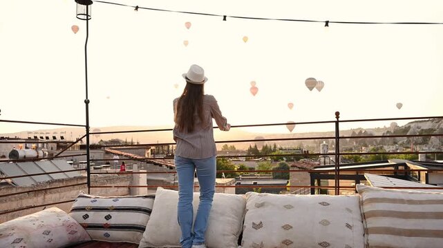Woman stands on hotel rooftop observation deck with patterned pillows and woven rugs on terrace, watching hot air balloon fly over city at sunrise in Cappadocia, Turkey. Tourism and ballooning
