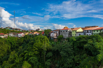 Fototapeta premium Cluster of vibrant houses perched on a lush green hillside with dense tropical foliage below, set against a vivid blue sky and scattered clouds in rural Phou Khoun, Laos. The scene highlights the