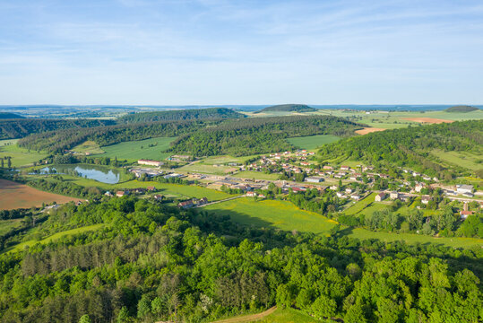 Aerial perspective of Varzy village surrounded by rolling green hills, dense forests, and a reflective lake under a clear spring sky, capturing vibrant rural scenery and patchwork fields.