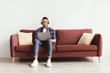 Obraz premium A man is sitting on a red couch with two cushions. He is using a laptop and wearing headphones. The room has a clean design with light walls and wooden floor.