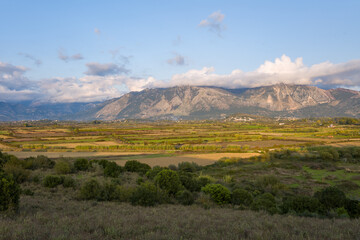 Obraz premium Layered agricultural fields stretch across a broad plain toward rugged Greek mountains partly covered by drifting clouds. Soft afternoon sunlight highlights the varied textures and green patches in