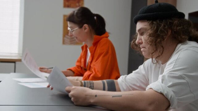 Side view shot of tattooed young woman as female inmate attending prison education program and reading tasks while sitting at desk in classroom, copy space