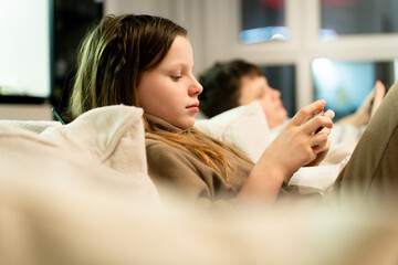 Schoolchildren at a cozy home on a sofa, focused on their smartphones playing mobile games. Scene captures modern childhood and the growing issue of phone addiction, digital dependency