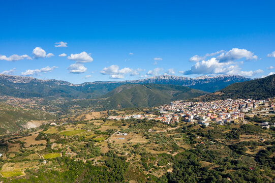 Aerial view of Orgosolo village surrounded by lush green hills and dramatic mountain ridges beneath a clear blue sky with scattered white clouds in Sardinia.
