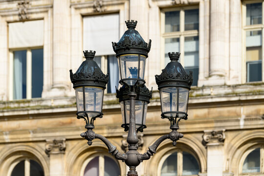 Elegant wrought iron street lamp featuring decorative crown details stands in front of the historic stone facade and arched windows of Hotel de Ville, illuminated by warm daylight.