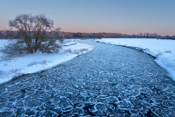 Beautiful nature landscape of winter river with cracked ice at sunset. Warm golden light reflects off the frozen surface, with a dark forest silhouette on the horizon.