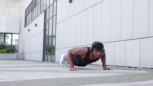 African American man in sportswear doing push-ups and exercising outdoors, athletic guy doing exercises with headphones on urban background