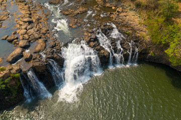 Sweeping aerial perspective of Tad Lo waterfall as water cascades over rugged rocks into a wide pool in southern Laos. Sunlit textures highlight the contrast between flowing water, exposed stone, and © Florent