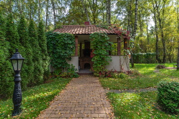 A path made of stones leads to a house surrounded by trees and plants. Vines cover the walls, and the ground is covered with fallen leaves. The scene shows the beauty of autumn