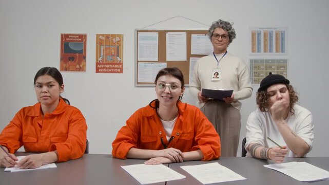 Front view shot of group of young women as female inmates sitting at desks and smiling at camera with grey haired female teacher in background during class in prison education program, copy space