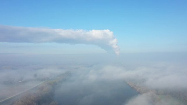 Aerial shot of the CNPE de Dampierre nuclear power plant emitting steam above a river, surrounded by morning fog and a clear blue sky in France.