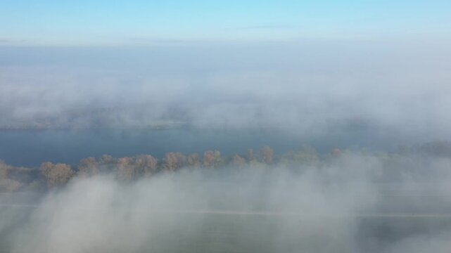 Aerial view of the Loiret countryside with dense morning fog partially covering a river and autumn trees. Tranquil rural landscape under a blue sky.