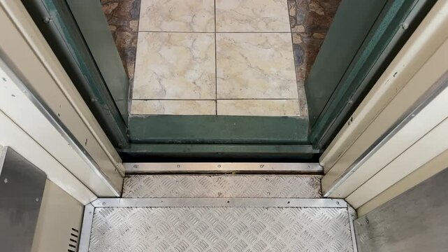 Point of view shot from inside an old elevator while the doors open. The metal cab floor contrasts with the building's marble tiles