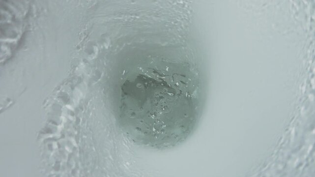 Close-up top view inside toilet bowl during flush. Water forms strong swirling vortex, moving toward drain hole. Hygiene concept, bathroom plumbing system in action, slow motion shot
