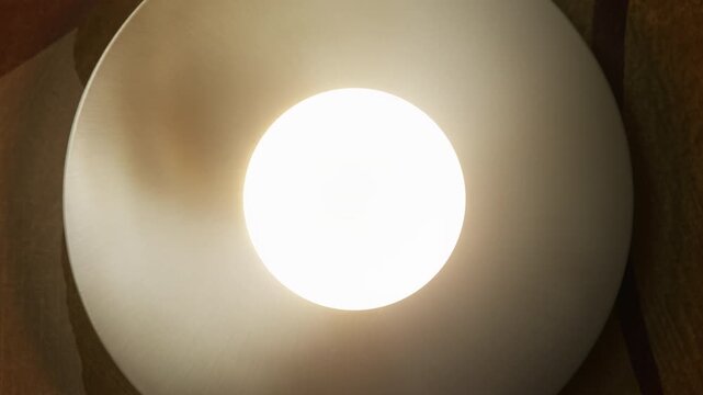 Centered shot of modern round steel wall lamp mounted on the wall. In dim evening light the room is barely visible, then soft warm glow turns on, highlighting circular metal form and smooth surface