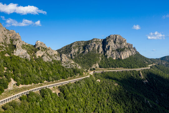 Elevated viaduct snakes along a lush green mountainside near Baunei and Dorgali in Sardinia, with dramatic rocky peaks rising under a clear blue sky.