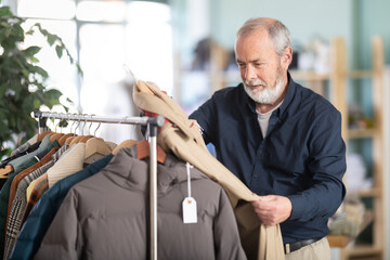 Senior man client pre-examine fabric and style of product, is busy choosing male trench. Client enjoys shopping, clothing and accessories for whole family, seasonal sale