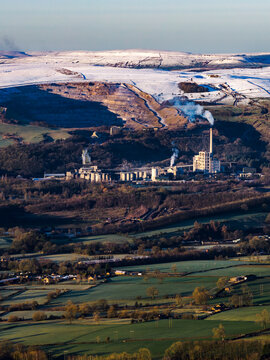 Aerial view of a factory emitting smoke nestled amidst the rolling hills and snow-dusted peaks of the Peak District, Buxton, Peak District National Park, United Kingdom.