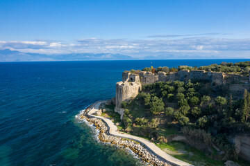 Naklejka premium Ancient stone ramparts overlook the deep blue Mediterranean, bordered by lush olive groves and a winding coastal path under a bright sky near Koroni, Greece.
