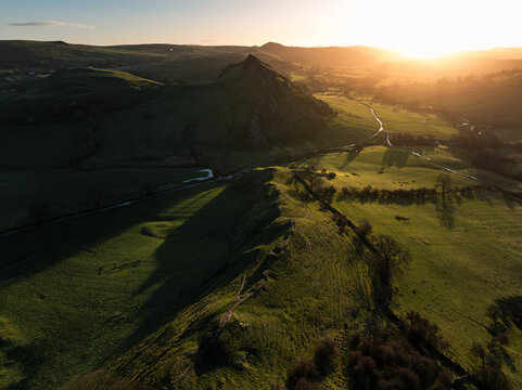 Aerial view of a sunlit ridge casting long shadows over the verdant fields of the Peak District, with a river snaking through the valley, Peak District, Peak District National Park, United Kingdom.