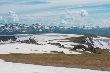 Scenic alpine landscape with sunlit snowy field on stony hill with view to big mountain range with forest and snow far away under cloudy sky. Stone outcrops among snows in sunlight in high mountains. © Daniil