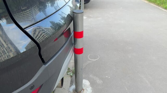 Crossover bumper scratched by metal pole, closeup rear bumper scuff on dark suv, parking edge near curb, red reflective post with paint transfer, urban outdoor light, gritty pavement.
