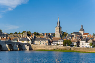 Naklejka premium Distinctive church spire rises above the old town skyline of La Charite sur Loire, with stone bridge arches and traditional houses lining the Loire riverbank. Bright blue sky and clear daylight