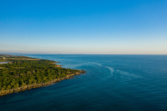 Aerial view of lush green coastline meeting the calm blue waters of the Ionian Sea near Arkoudi, Greece. The scene features clear sky, rocky shore, and gentle sunlight creating a tranquil
