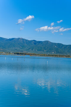 Still blue water of a salt lagoon reflects scattered clouds beneath a vivid sky, with a line of green mountains in the distance. Subtle poles in the water and gentle ripples evoke a peaceful wetland