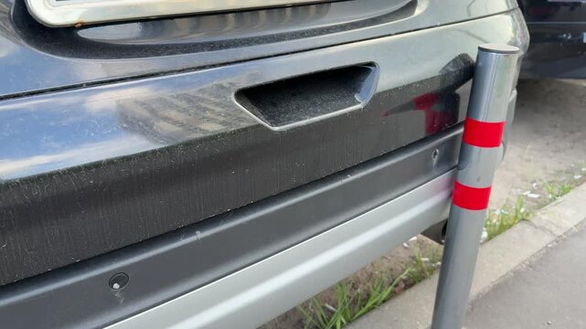 Rear bumper scratched by bollard, black crossover parked tight against reflective post with red tape, paint scuff along lower silver trim, closeup curbside shot showing minor damage.