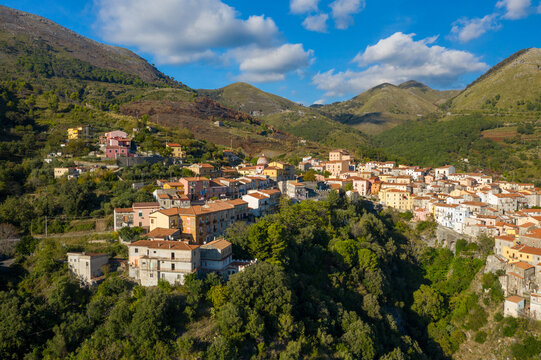 Vibrant buildings with terracotta roofs line the steep hillside of Tortora, Italy, surrounded by lush greenery and dramatic mountain peaks under a bright blue sky with scattered clouds.