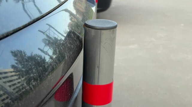 Rear bumper scratched by bollard, black crossover parked tight against reflective post with red tape, paint scuff along lower silver trim, closeup curbside shot showing minor damage.