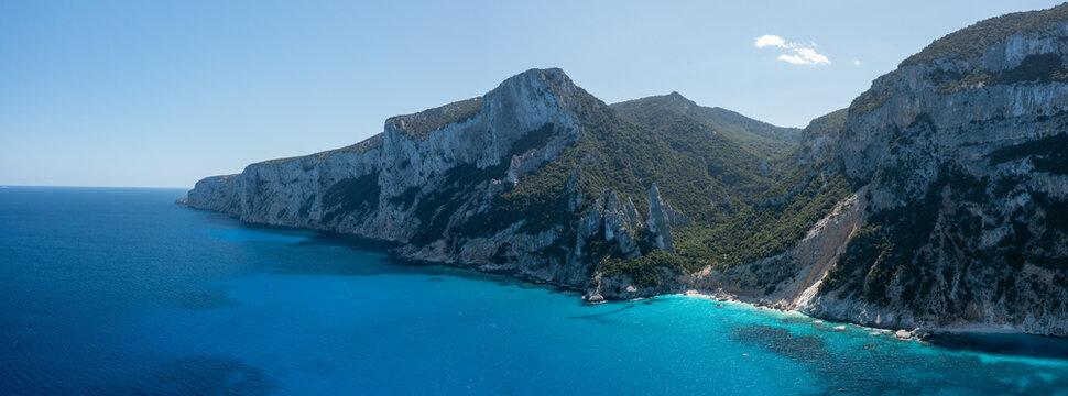 Expansive view of steep limestone cliffs meeting vibrant azure waters at Cala Goloritze, Baunei, Sardinia. The rugged coastline contrasts with the deep blue Mediterranean under clear daylight.