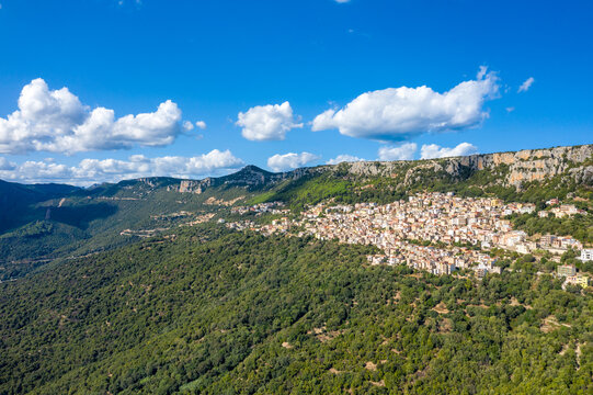 Aerial view of Baunei perched on a densely forested slope beneath towering limestone cliffs in Sardinia, Italy. Bright sunlight and scattered clouds create a vivid Mediterranean atmosphere.