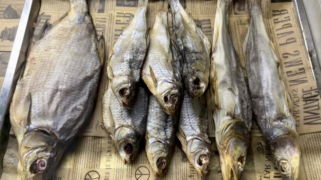Dried fish lined on newsprint counter, salted heads arranged in tight row with golden gray scales and flaky brittle texture, closeup rustic display suggesting tavern snack sale for beer.