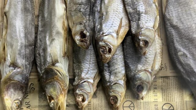 Dried fish lined on newsprint counter, salted heads arranged in tight row with golden gray scales and flaky brittle texture, closeup rustic display suggesting tavern snack sale for beer.