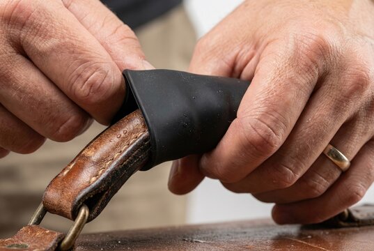 A person's hands stretch a new black rubber grip cover over the worn brown leather handle of a vintage suitcase to repair and improve it.