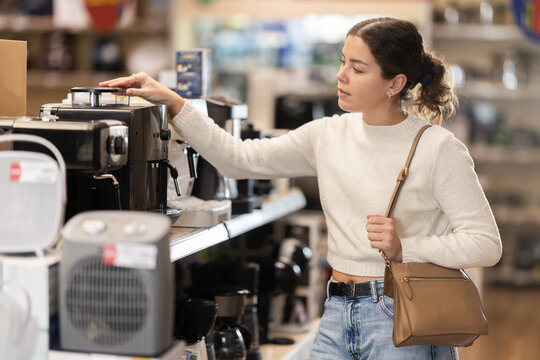 Girl buyer examines coffee machine in detail before buying, checks completeness of household appliances and presence of defects. Customer chooses household appliances in store exhibition hall.