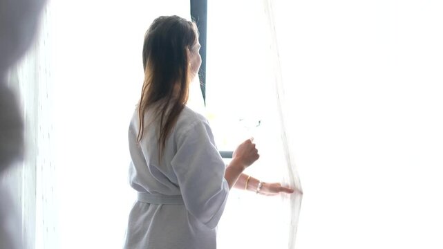 girl with a glass of water dances by the window in a white robe woman smiling and opening curtains to let morning sunlight into the room fresh new day, hope, positive mindset and peaceful lifestyle.
